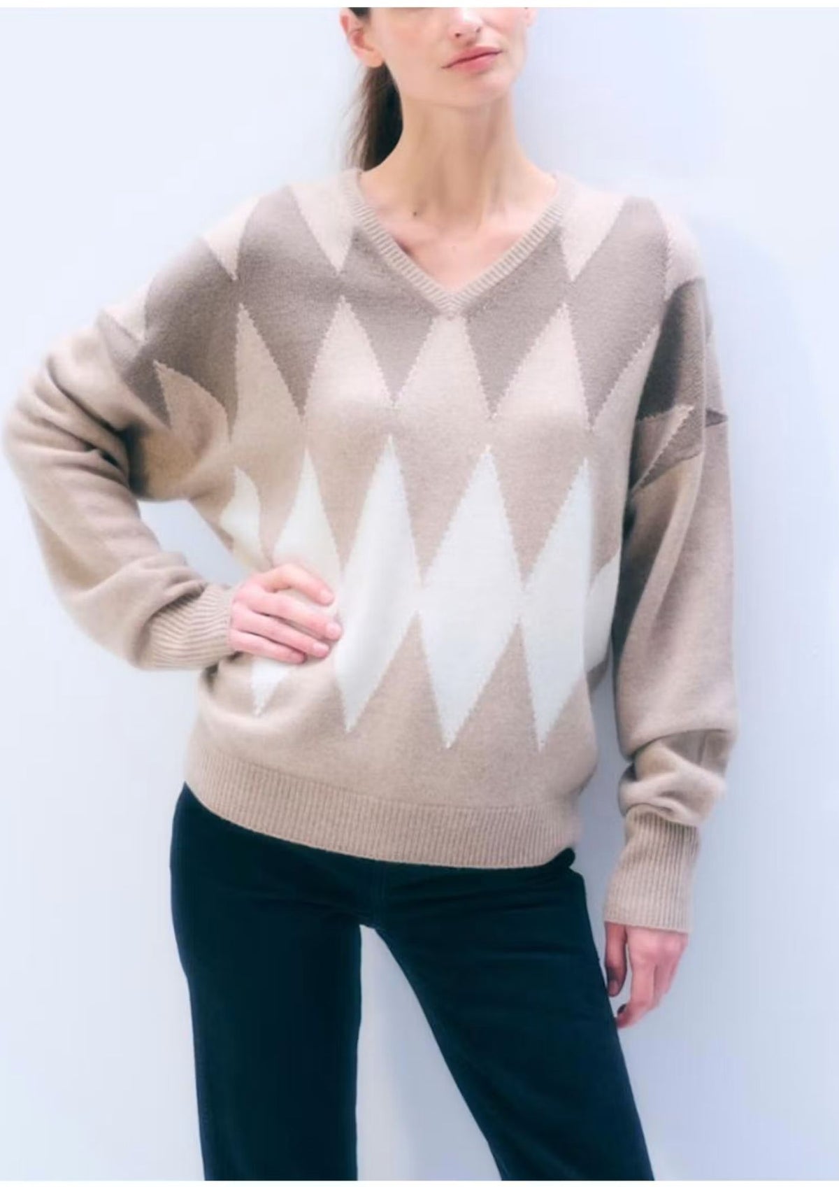 A woman wearing a beige and cream argyle knit sweater with a V-neck and dark pants, standing against a light gray background.