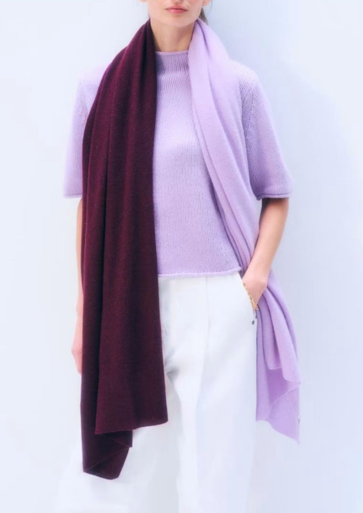 A model wearing a light purple knit sweater and white trousers, styled with a dual-color scarf draped over the shoulders in deep burgundy and light purple.