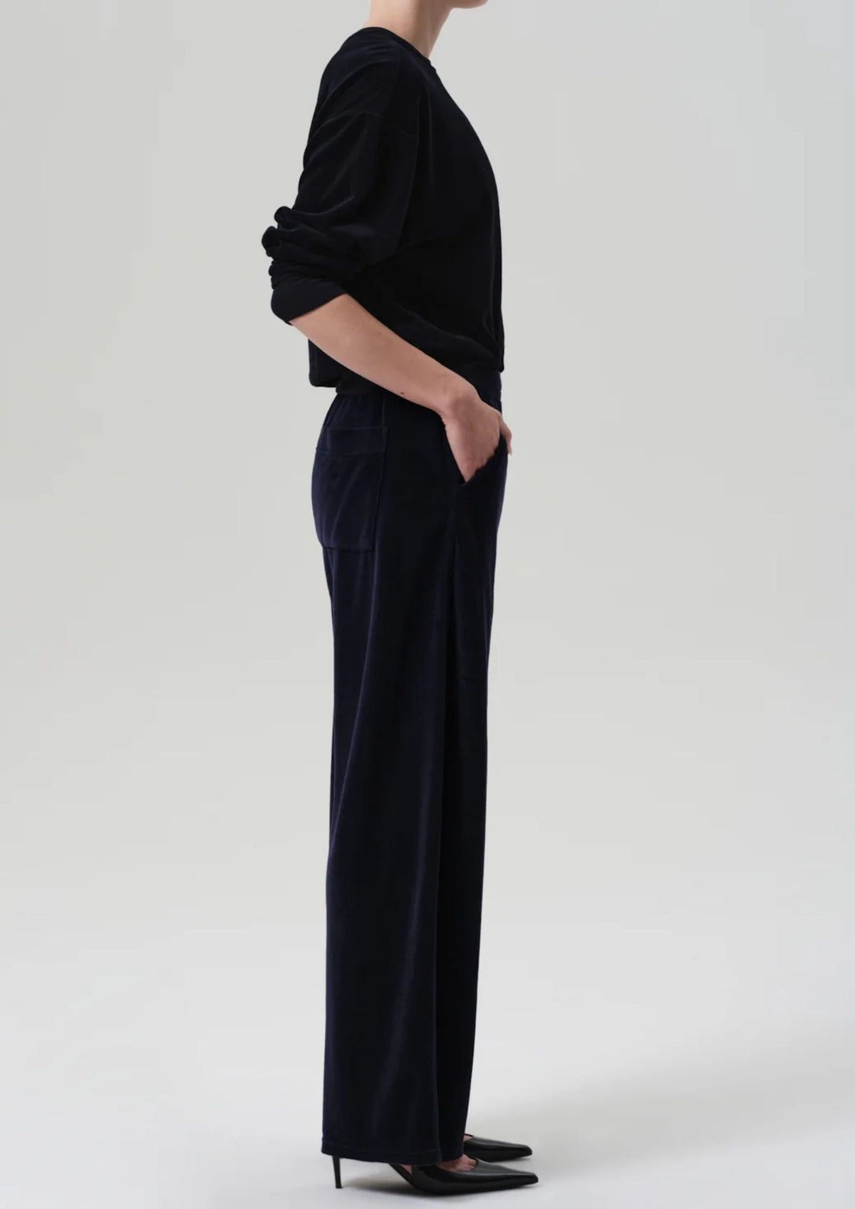 A side-profile view of a model wearing a black velvet long-sleeve top and matching wide-leg trousers, paired with black pointed-toe heels, against a light gray studio background.