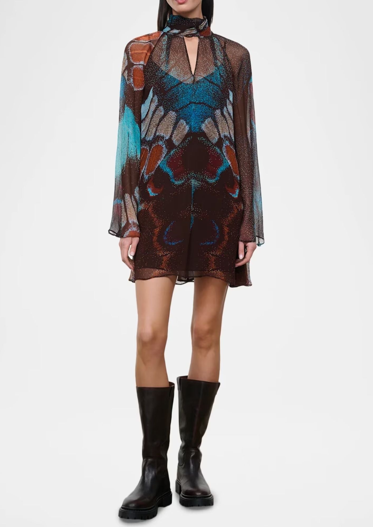A model wearing a sheer, long-sleeved mini dress with a complex abstract butterfly-like pattern in brown, teal, and rust, paired with black knee-high boots against a plain white studio background.