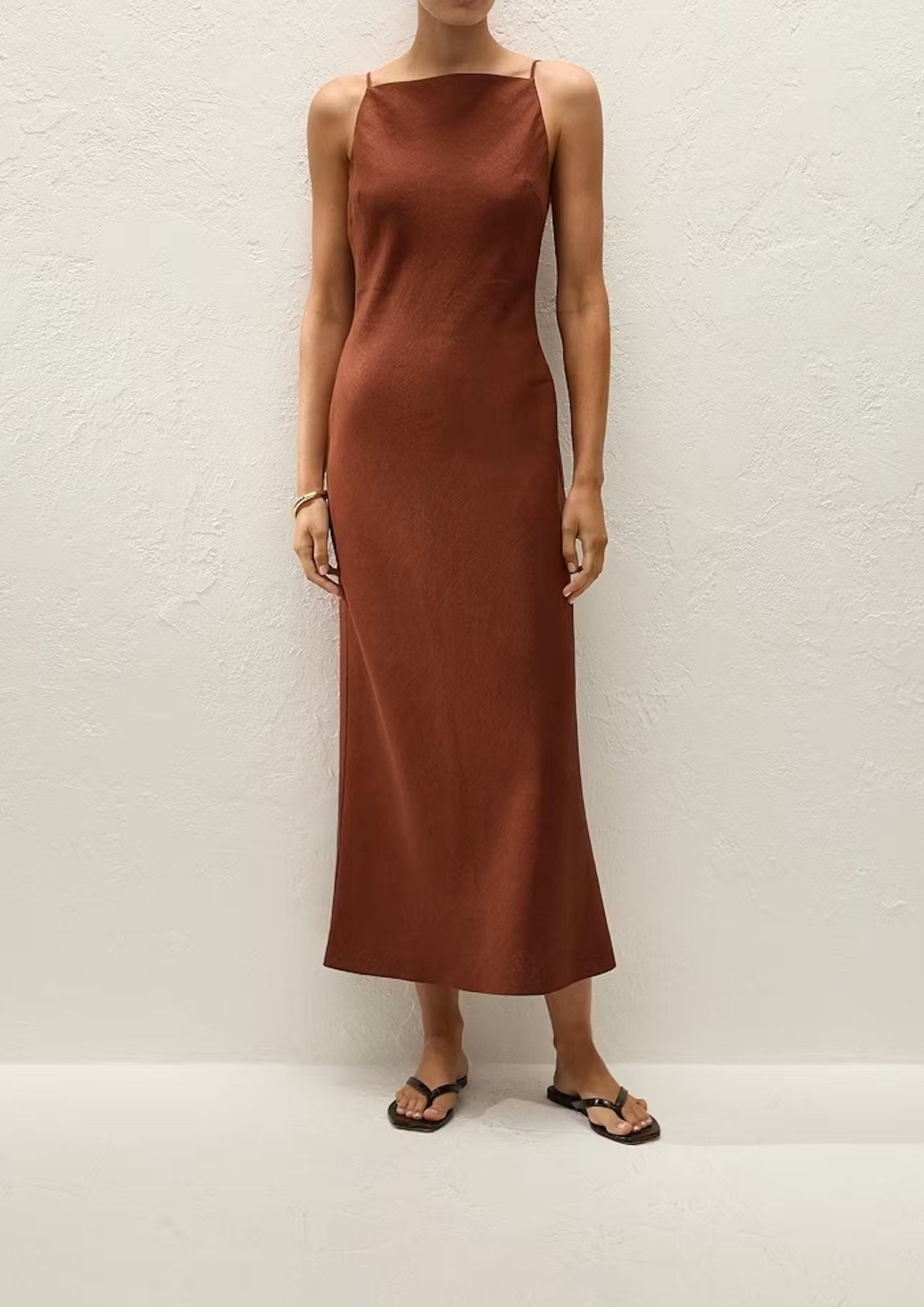 A woman modeling a sleeveless, high-neck maxi dress in a warm rust brown color, standing against a textured white wall.