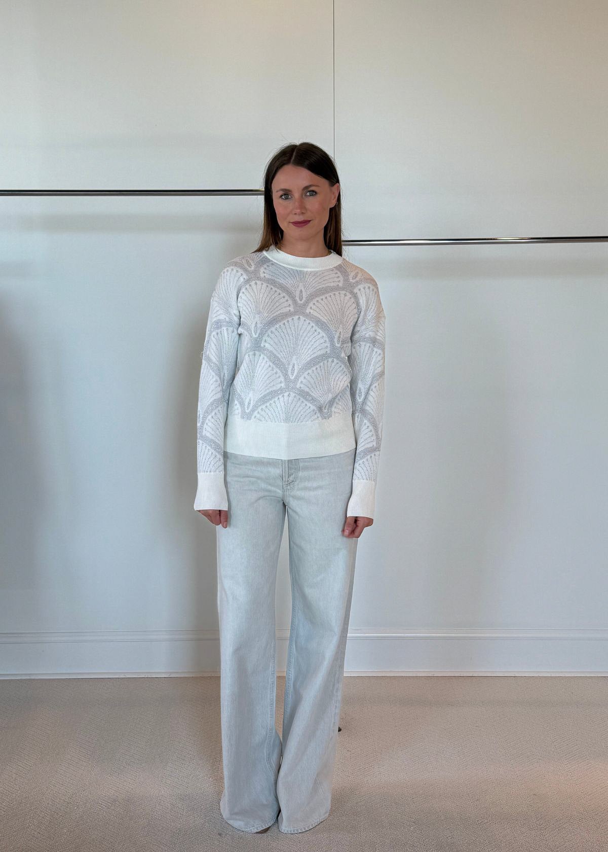 Woman wearing a white patterned sweater and light gray pants against a plain background
