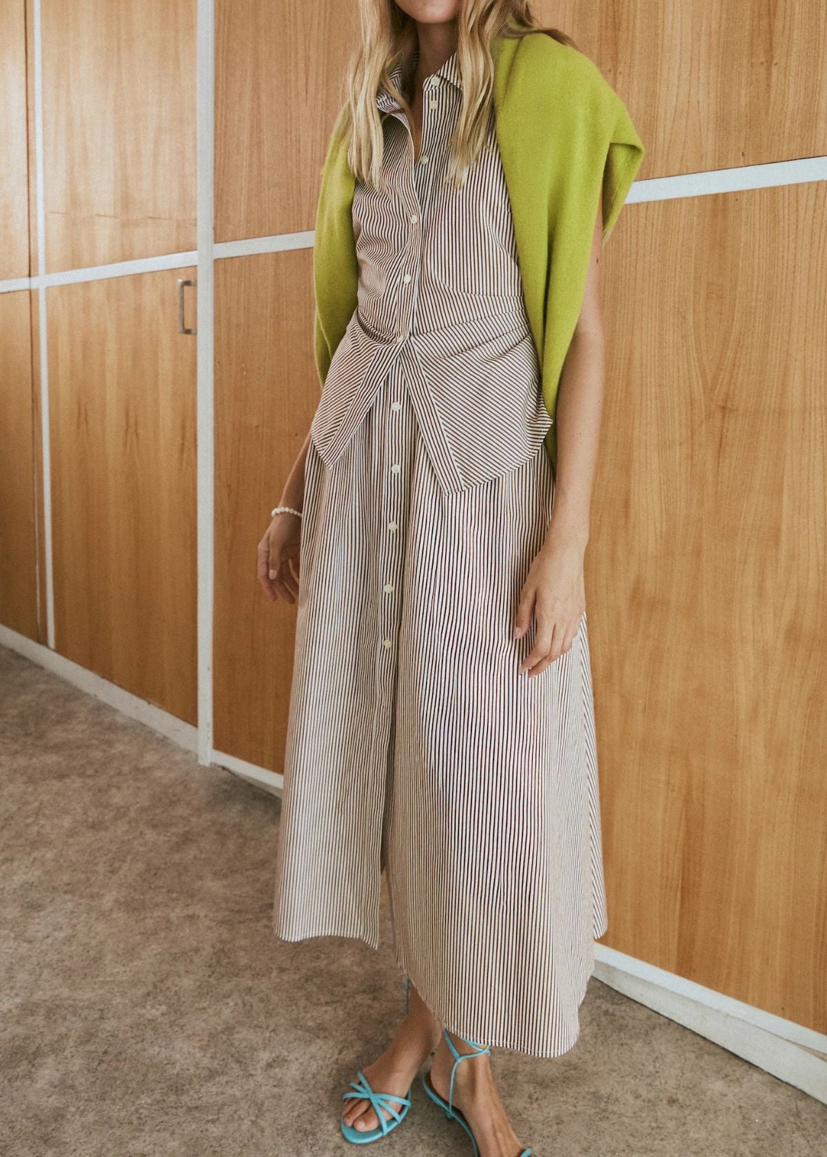 Woman wearing a long striped dress with a green cardigan in a room with wooden walls.